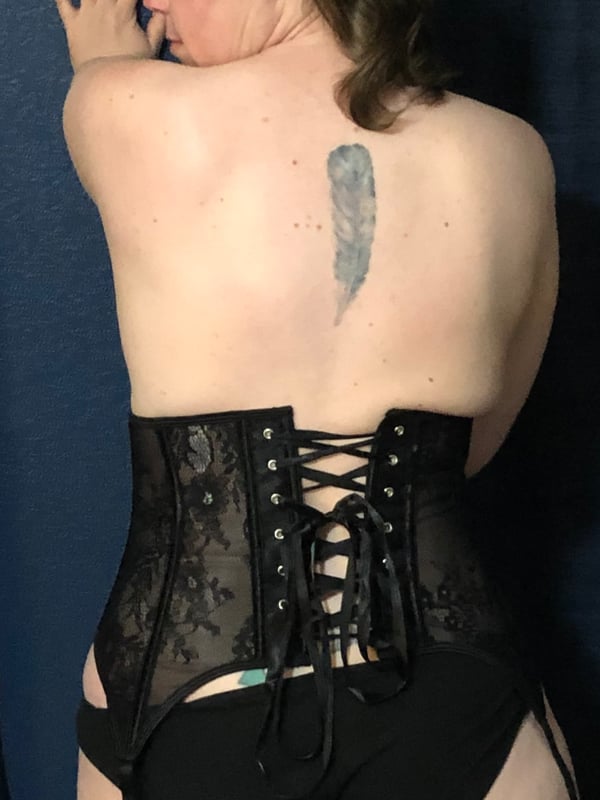 Woman from behind wearing a black lace corset and bottoms, posing against a blue curtain with a feather tattoo on her upper back.