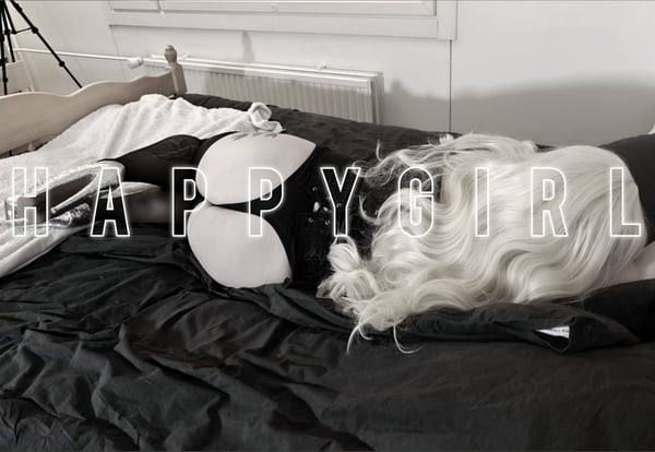 A person lying on a bed with long blonde hair, wearing a black top and white bra cups, in a cozy indoor setting with a tripod nearby.