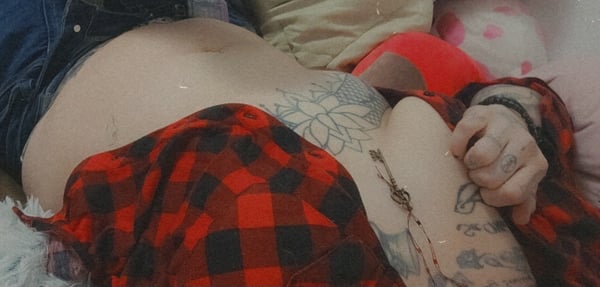 Person reclining on a bed with pillows, wearing plaid shirt and jewelry, displaying tattoos on torso.