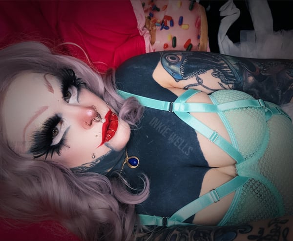 A woman with heavy tattoos and piercings reclines on a bed wearing a green harness top, surrounded by colorful pillows and blankets.