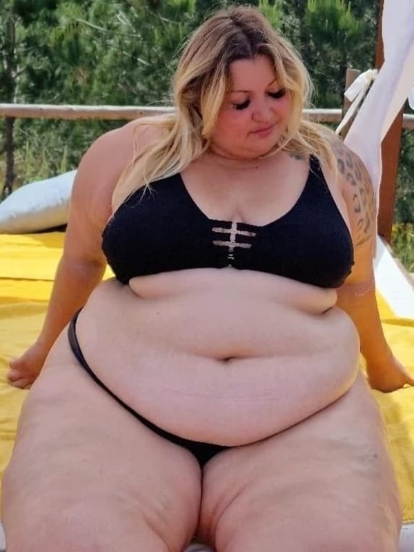 A woman with blonde hair and tattoos sitting on a yellow lounge chair by a poolside, dressed in a black bikini top and bottoms.