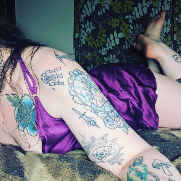 A woman with extensive tattoos lounges on a bed in a silky purple slip, surrounded by patterned decor.
