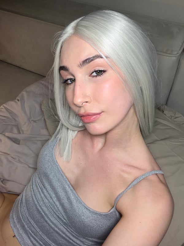 A close-up of a person with platinum hair wearing a gray top, resting on a bed.