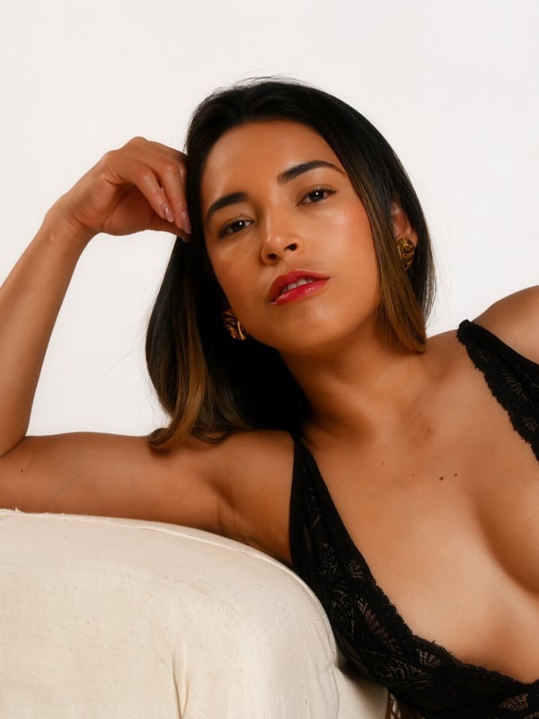 A woman with long wavy dark hair posing thoughtfully on a white surface, wearing a lace top, with a photography studio logo in the corner.
