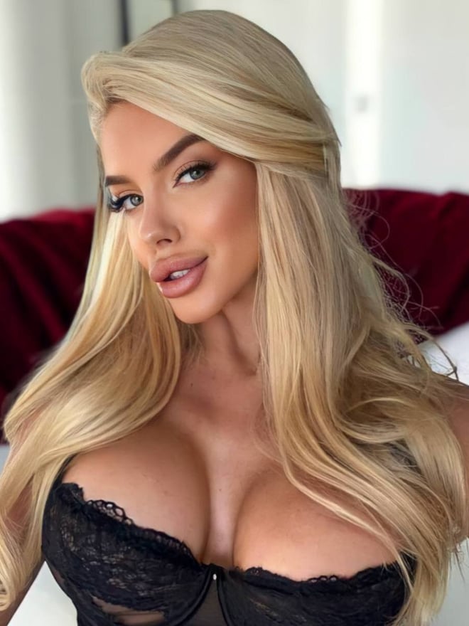 A close-up portrait of a blonde woman with long hair wearing a black lace top.