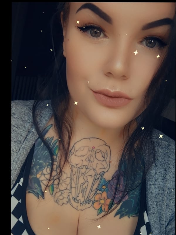 Close-up portrait of a woman with bold eyebrows, dark hair, and extensive neck tattoos, wearing a gray blazer.