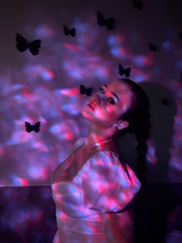 A young woman with brunette hair in a ponytail poses with her head tilted in a purple-lit room decorated with black butterfly cutouts, wearing a light off-shoulder top.
