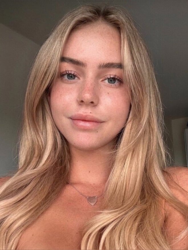 Close-up portrait of a young woman with long blonde hair, subtle makeup, and a necklace, smiling softly against a plain background.