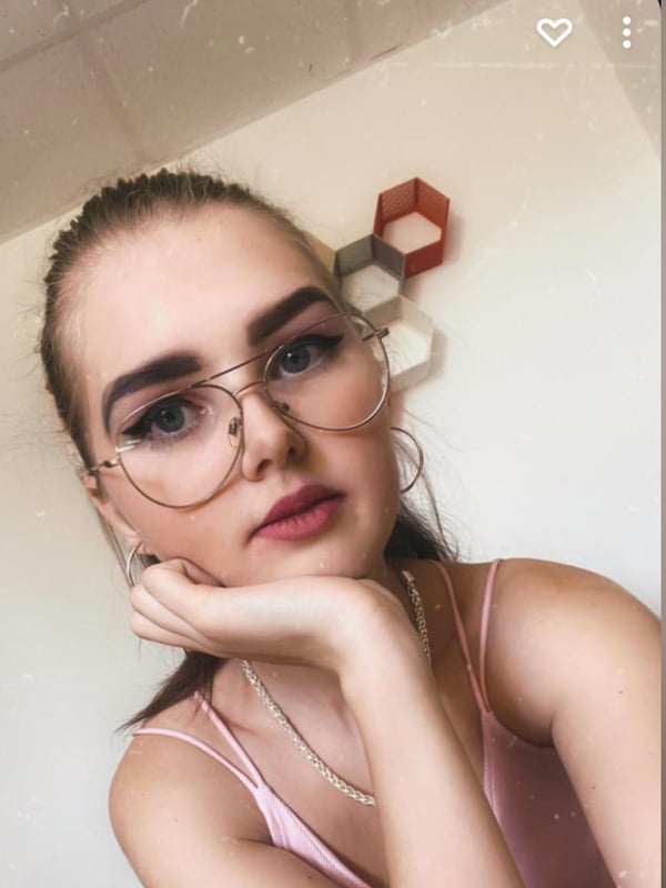 A young woman with glasses and ponytail resting her chin on her hand, wearing a pink top in a casual indoor setting.
