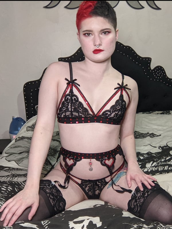 A woman with split-colored hair sitting on a bed in black lace lingerie and stockings, posed against a dark gothic bedroom background.