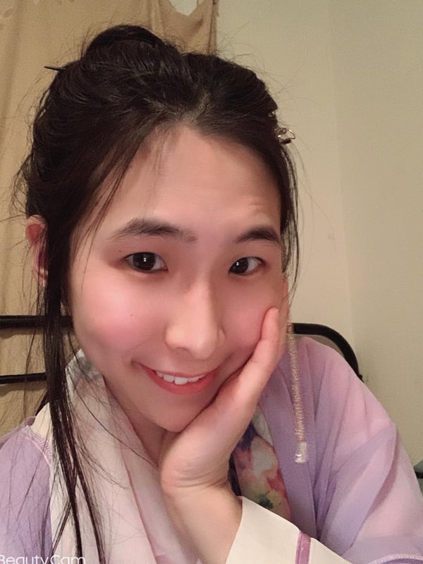 Young woman with dark hair in a bun smiling with hand on cheek, wearing a light purple top in a simple indoor setting.