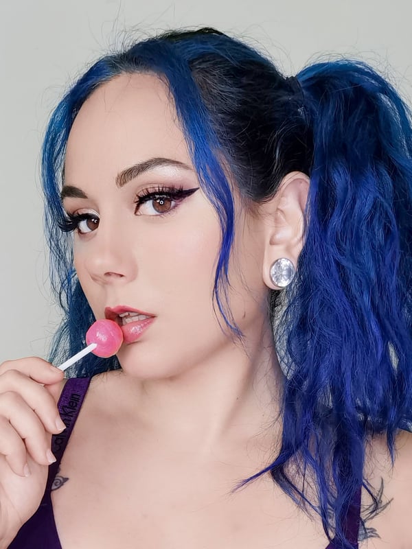 A woman with blue dyed hair in pigtails, smiling while holding and licking a lollipop, wearing hoop earrings and a top.
