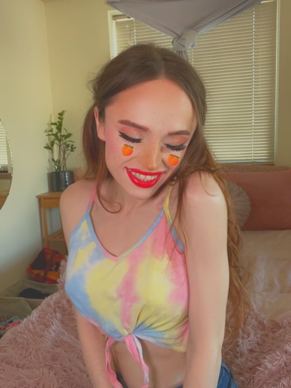 Young woman with long brown hair and freckles, wearing colorful makeup and a tie-dye tank top, posing playfully on a bed in a softly lit bedroom.