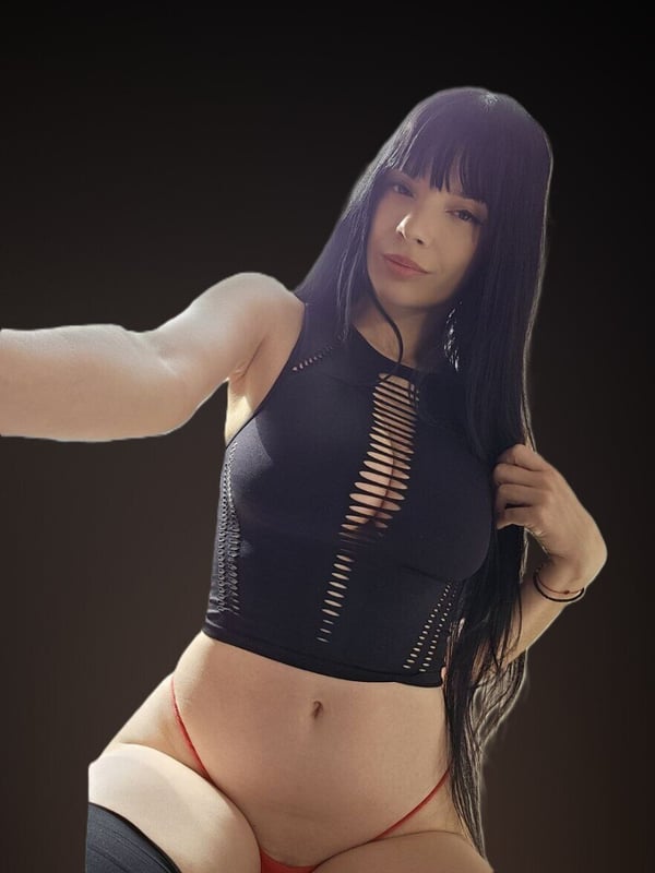 A woman with long dark hair posing confidently in a black mesh top and red bottoms against a dark background.
