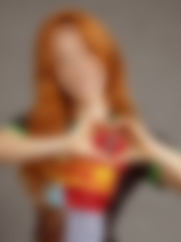 Close-up of a young woman with long red hair making a heart shape with her hands, wearing a colorful top, against a gray background.