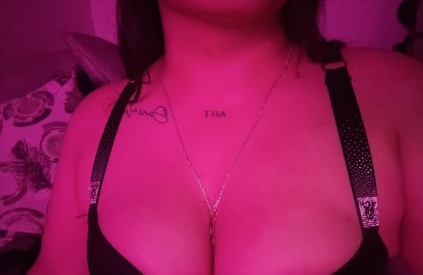Woman posing in a dimly lit room with pink lighting, wearing a black top and necklace with jewelry.