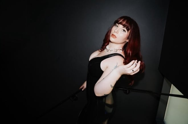 A woman with long red hair posing confidently in a black dress against a dark background with stairs.