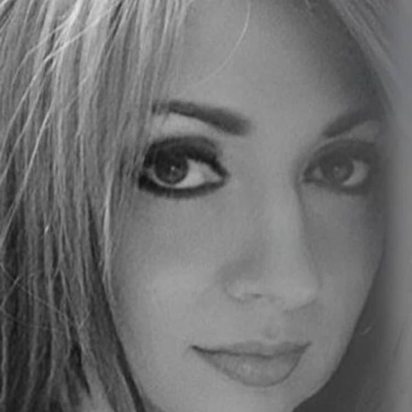 Close-up portrait of a woman with long blonde hair, dramatic makeup, and a confident smile in black and white.