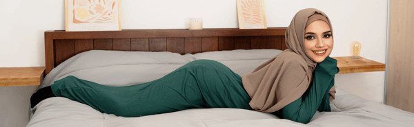 A woman in a hijab and long modest clothing poses playfully on a bed with artwork on the wall and a candle nearby.