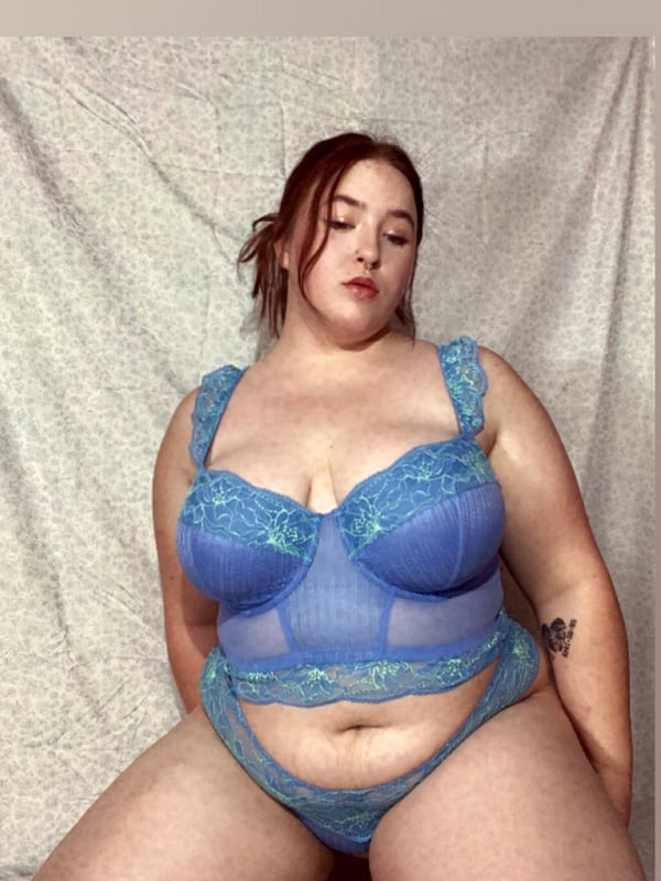 A woman with red hair posing on the floor in a blue lace lingerie set, reclining against a gray background.