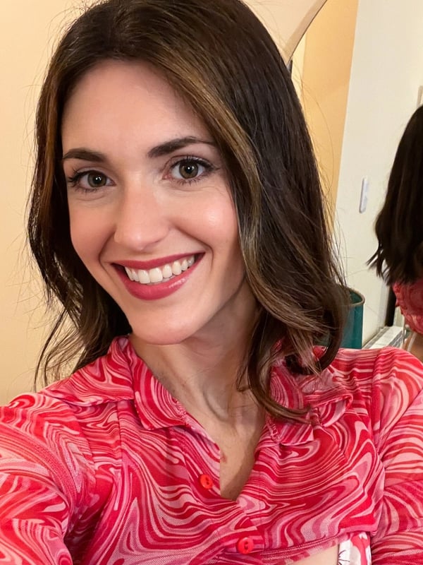 A smiling woman with long brown hair wearing a sheer red top, standing in a room with a mirror reflection and indoor plants.
