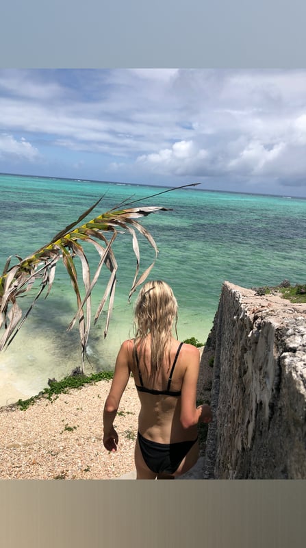A woman with long blonde hair stands by a tropical beach, wearing a black bikini top and bottoms, with turquoise water and palm trees in the background.