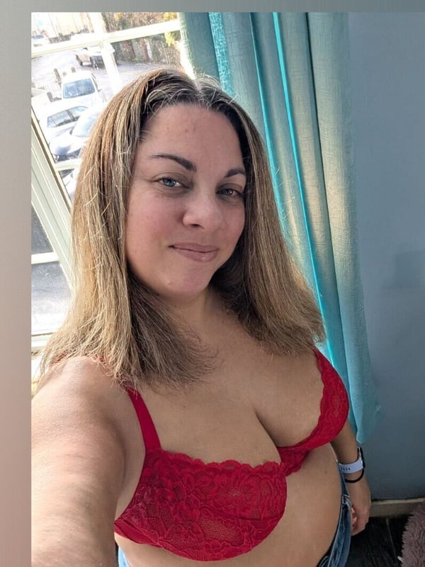 A smiling woman taking a selfie in a red lace top by a window with curtains and a view outside.