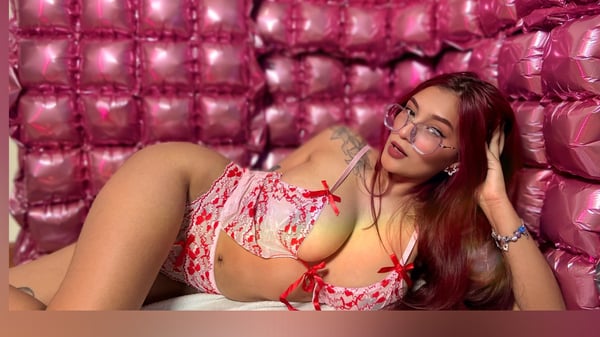 A woman with glasses and long red hair reclining on a bed against a pink balloon wall, wearing lingerie and smiling playfully.