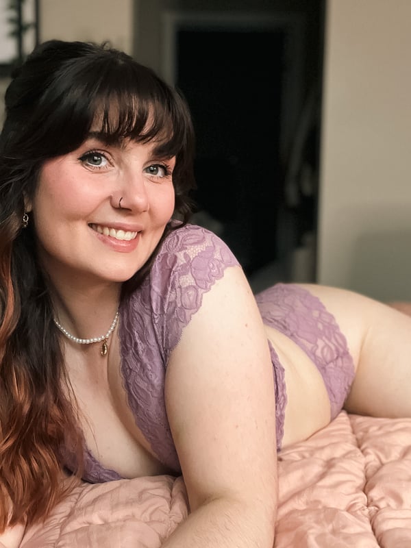 A smiling woman with long dark hair lounging on a bed in lace attire inside a bedroom.