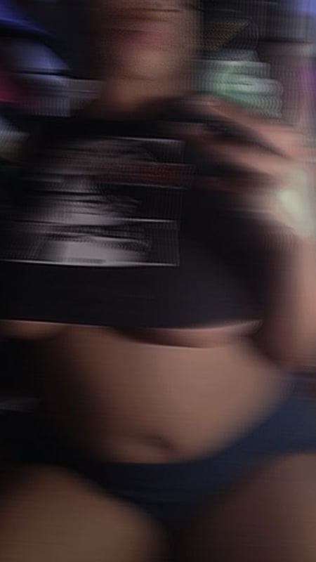 A person taking a close-up selfie in a dimly lit room, wearing a dark top and shorts, with a phone screen visible.