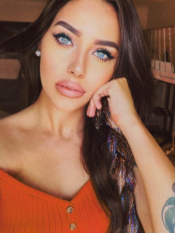 Close-up portrait of a woman with long brunette hair, blue eyes, and subtle makeup, posing with her hand on her face in a cozy indoor setting.