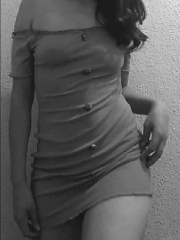 A woman standing in a form-fitting off-shoulder dress against a plain wall, captured in black and white.