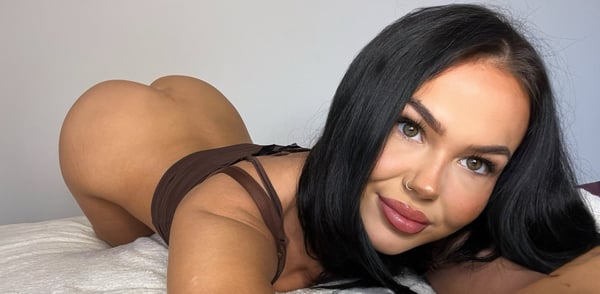 A woman with long dark hair posing on a bed in a brown outfit against a plain background.