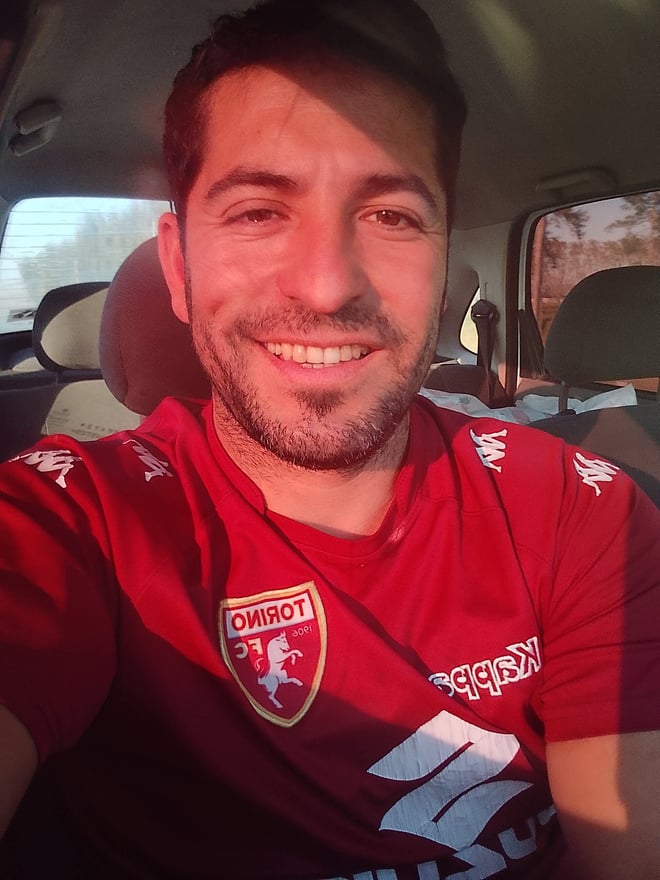 Man smiling in a red sports jersey, seated in a car.