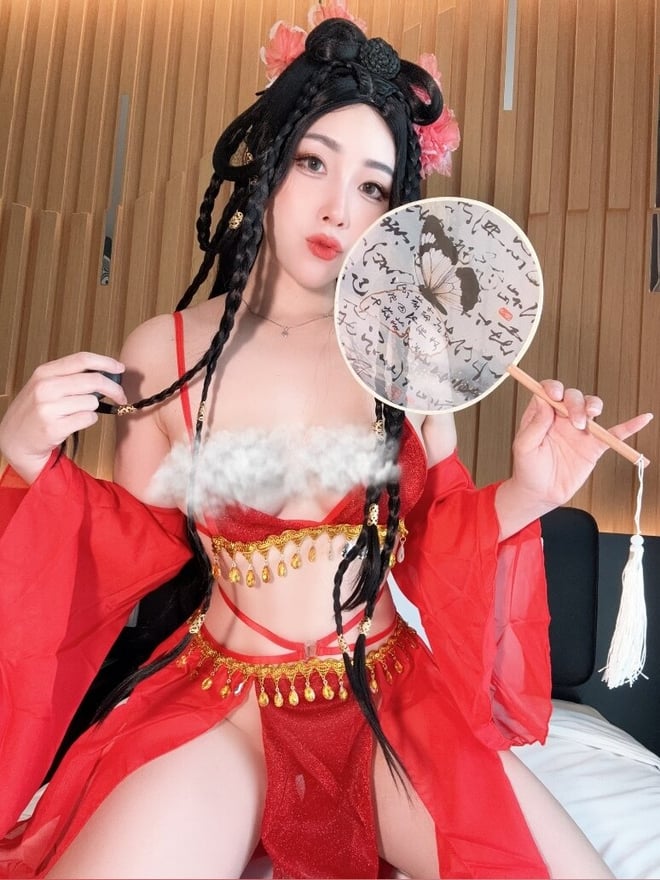 A woman in an elaborate red traditional costume with floral hair accessories holding a fan, posing indoors with wooden paneling.