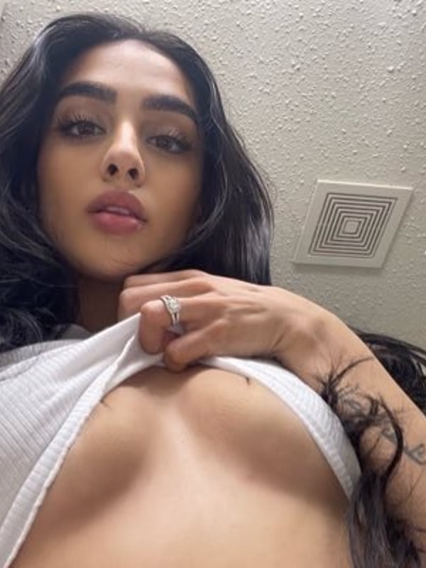 A woman taking a close-up selfie in a bathroom, wearing a white top and showing a ring on her finger.