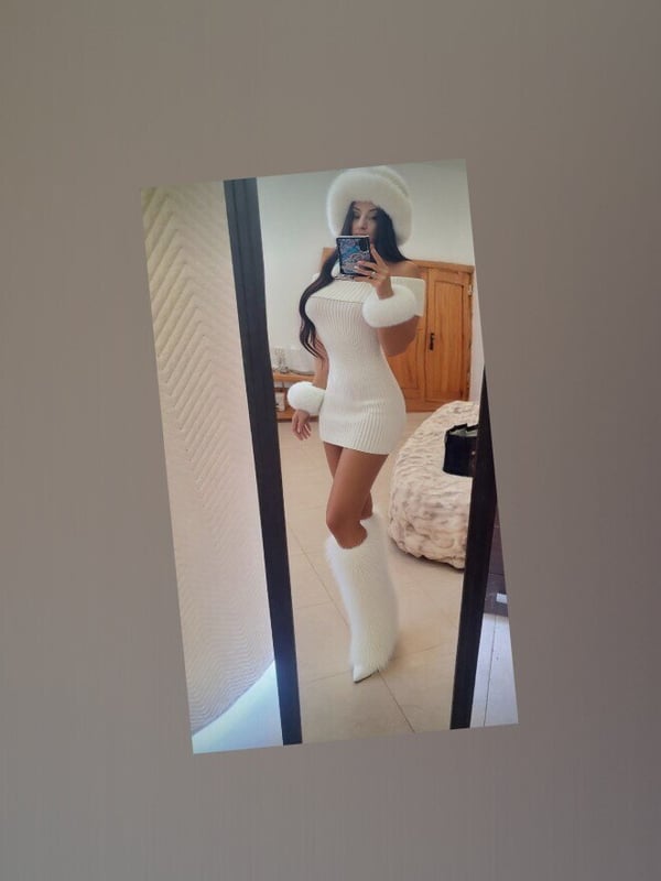 Woman in a white dress and boots posing in front of a mirror in a bedroom with wooden furniture.