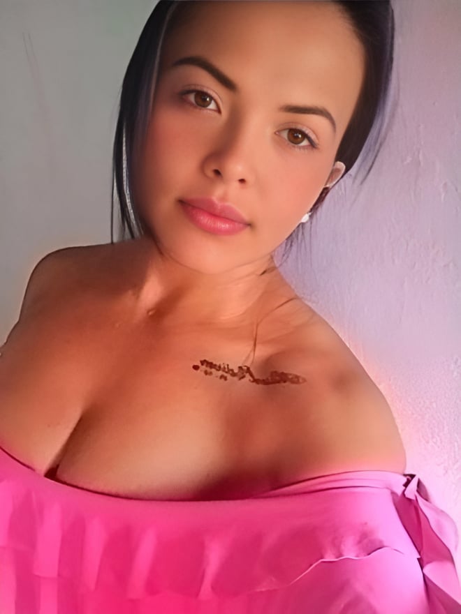 A woman with long dark hair and subtle tattoos on her shoulder smiles at the camera, wearing a pink off-shoulder top.