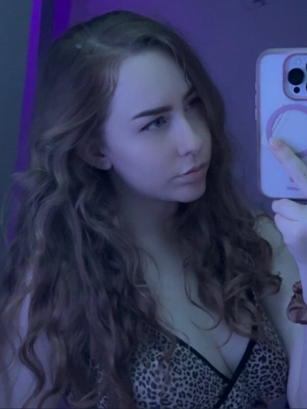A young woman taking a mirror selfie in a purple-lit bathroom, holding her phone and wearing leopard print lingerie.