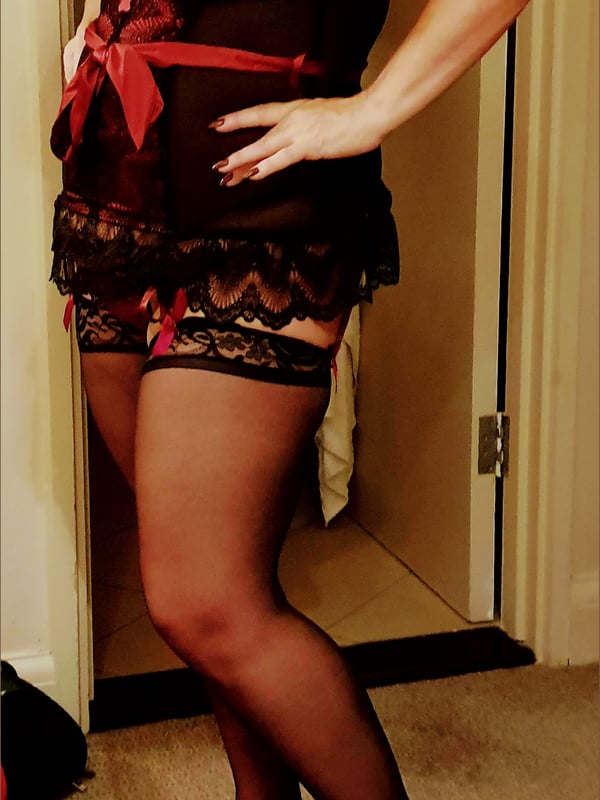 Older woman posing in a red lace lingerie top and black stockings near a door.
