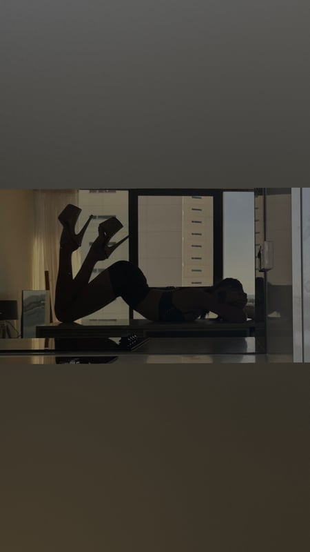 Silhouette of a woman reclining on furniture near a window with high heels, against a cityscape view.