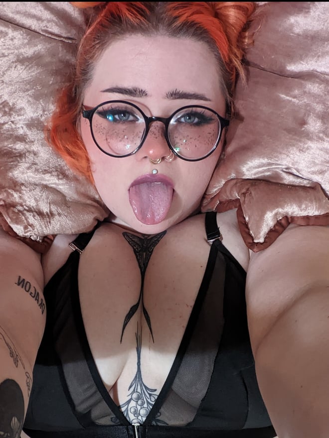A person with orange hair and glasses lying on pink pillows, wearing a black top, sticking out their tongue with visible piercings.