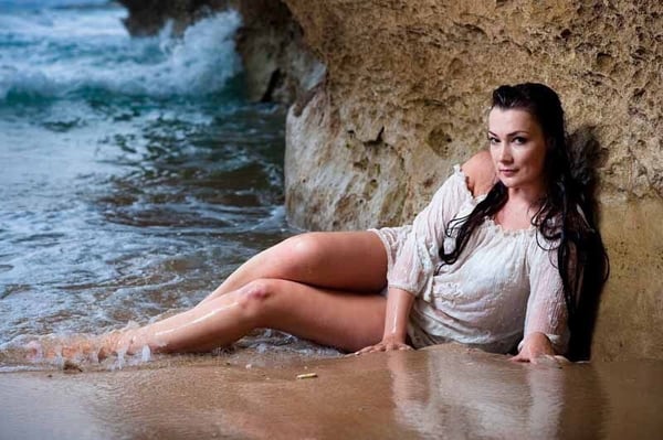 Woman with long dark hair lounging on a sandy beach near ocean waves and rocky cliffs, wearing a light off-shoulder dress.