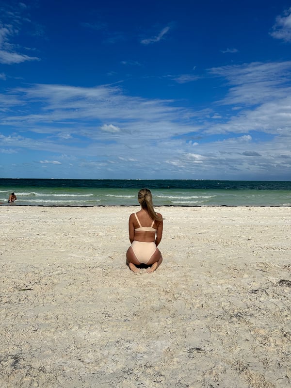 A young woman with long blonde hair sits on a sandy beach facing the turquoise ocean under a blue sky, wearing a light-colored bikini top and bottoms.