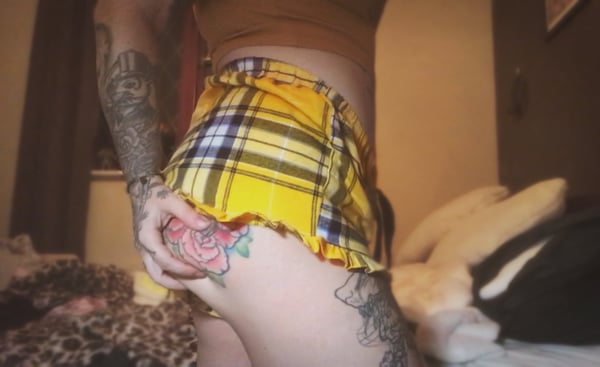 A woman posing in a plaid skirt and top, showcasing tattoos on her arms and leg in a cozy indoor setting with pillows.