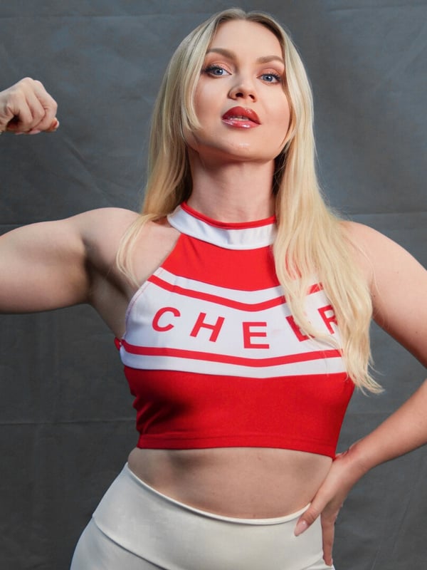 A blonde woman flexing her arm muscles while wearing a red and white cheerleader top and white pants against a gray background.