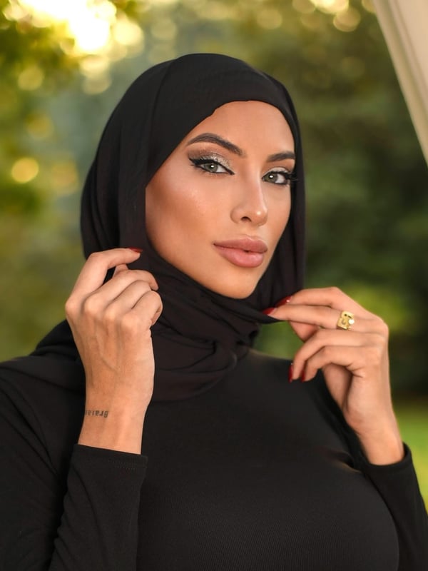 A woman wearing a black hijab and dress, posing with hands adjusting her hijab against a blurred outdoor background.