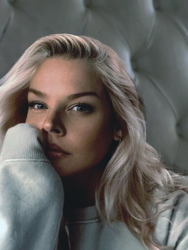 A woman with long blonde hair posing thoughtfully in a light sweater against a gray upholstered background.