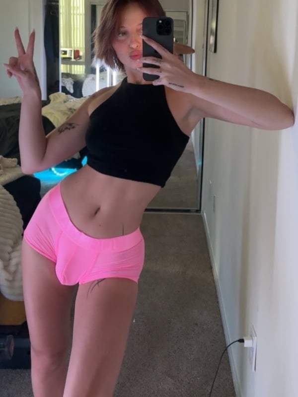 A person with short blonde hair wearing a white halter top, posing confidently in a purple-lit room.