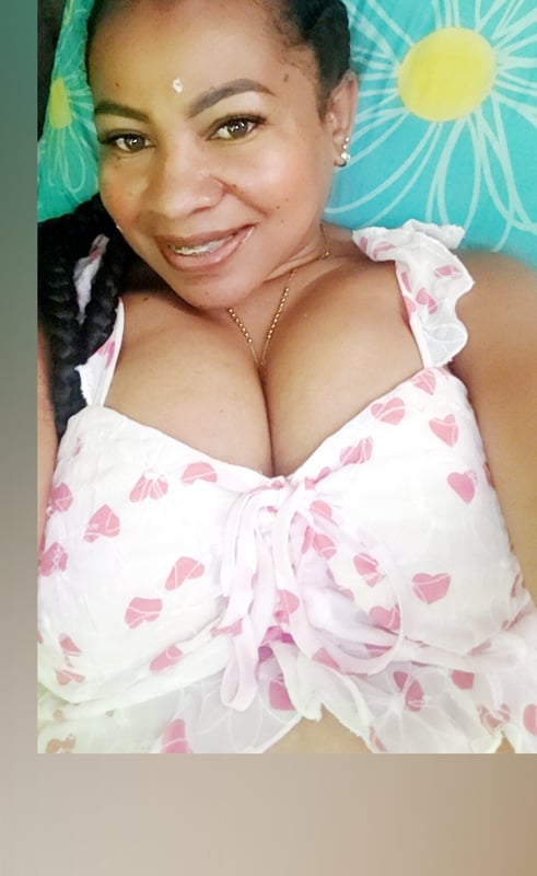 Woman with braided hair smiling while reclining on a bed with colorful floral pillows and a patterned top.